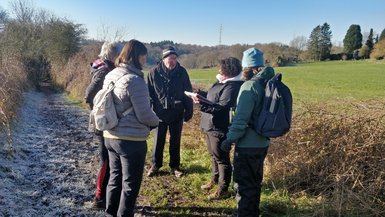 Les deux guides et les trois participants sont rassemblés en cercle pour discuter sur un chemin campagnard encore givré. Derrière, se trouvent les prairies baignées de soleil. L'image est moins zoomée et montre davantage le paysage.