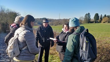 Les deux guides et les trois participants sont rassemblés en cercle pour discuter sur un chemin campagnard encore givré. Derrière, se trouvent les prairies baignées de soleil. L'image est centrée sur le groupe.