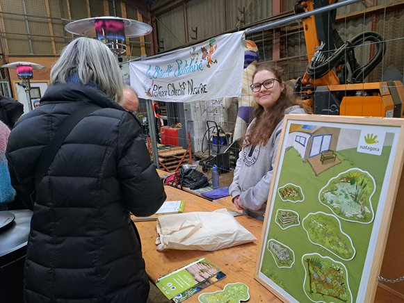 Une bénévole pour Natagora souritfièrement pour la photo. À gauche, devant elle, un couple de dos s'inscrit pour devenir membre Natagora. Un welcome pack leur est offert en remerciement. À droite, à l'avant-plan, on voit le jeu magnétique pour sensibiliser au jardin naturel.