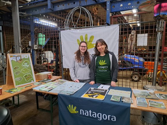 Deux femmes bénévoles pour Natagora dont celle de droite porte un t-shirt Natagora, sourient pour la photo en tenant le stand constitué d'une table sur laquelle se trouvent des flyers, livrets, posters, affiches, images, etc. portant sur la nature et les activités de Natagora. À gauche, un jeu magnétique pour conscientiser au jardin naturel. Derrière et devant elles se trouvent des banderoles avec le logo de Natagora en vert.