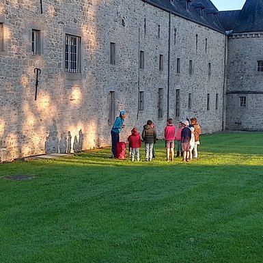 Photo d'une dizaine d'enfants debout écoutant une animatrice. Ils sont sur l'herbe, baignés dans les rayons du soleil du petit matin.