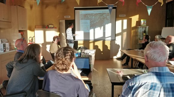 Photo de Frédéric faisant une présentation des réserves naturelles de Natagora devant un écran montrant une carte d'une réserve. Les participants, à l'avant plan, l'écoutent avec attention.