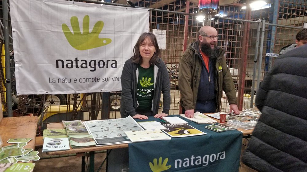Une femme portant le t-shirt de Natagora et un homme se tiennent derrière un stand constitué d'une table sur laquelle se trouvent des flyers, livrets, posters, affiches, images, etc. portant sur la nature et les activités de Natagora. Derrière eux, une banderole avec le logo de Natagora en vert.